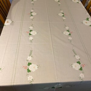 May include: A rectangular light gray tablecloth featuring a repeating pattern of white flowers with green leaves and small red accents arranged in vertical lines along the center and edges of the fabric surface.
