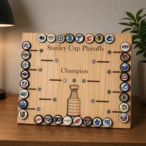 Op de afbeelding: Houten Stanley Cup Playoffs bord met teamlogo's. Het bord toont de tekst "Stanley Cup Playoffs" en "Champion", samen met een gegraveerde Stanley Cup trofee. Inclusief magnetische team pucks voor het volgen van de play-offs.