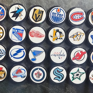 May include: A selection of hockey pucks, each displaying a different NHL team logo. The pucks are black with white circular faces, showcasing team emblems in various colours. Logos include the Carolina Hurricanes, New York Rangers, and Toronto Maple Leafs.