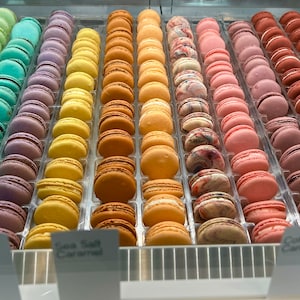 Peut inclure: Présentation de macarons colorés, disposés en rangées par couleur. Les macarons varient du vert au violet, jaune, orange, rose et marron. Des étiquettes telles que "Citron", "Caramel au sel marin" et "Barbe à papa" sont visibles.