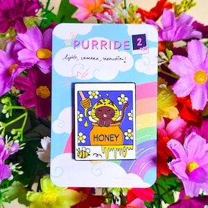 Peut inclure: Une épingle émaillée Purride avec un chat brun portant un chapeau d'abeille, assis dans un pot de miel. L'épingle est sur une carte bleu clair avec le texte "Purride" et "light, camera, meowtion!" entourée de fleurs et d'un arc-en-ciel.
