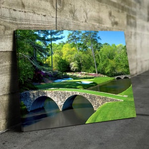 Puede incluir: Una vista panorámica de un campo de golf con un puente de piedra sobre un estanque. El césped verde es exuberante y los árboles están en plena floración. El puente está hecho de piedra y tiene dos arcos. El estanque está tranquilo y refleja el cielo.