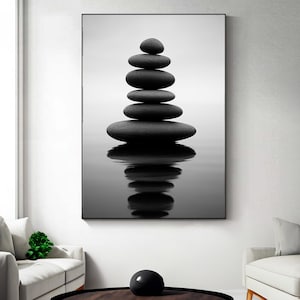 May include: Black and white photograph of a stack of smooth, dark grey stones balanced on top of each other. The stones are reflected in the water below, creating a symmetrical image. The artwork is displayed on a wall.