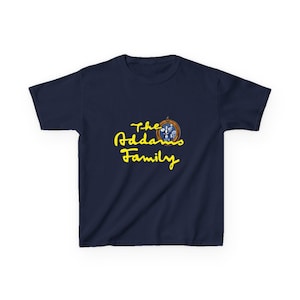 May include: A navy blue short-sleeved t-shirt featuring the yellow text The Addams Family in a cursive script. Above the text is a small, oval-shaped brown frame containing a graphic illustration of the Addams Family characters in blue and white.