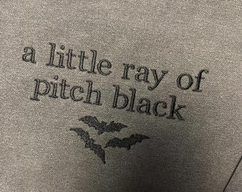 May include: A gray sweatshirt with the embroidered phrase "a little ray of pitch black" in black thread. Below the text are three bat silhouettes, also in black. The fabric has a textured appearance.