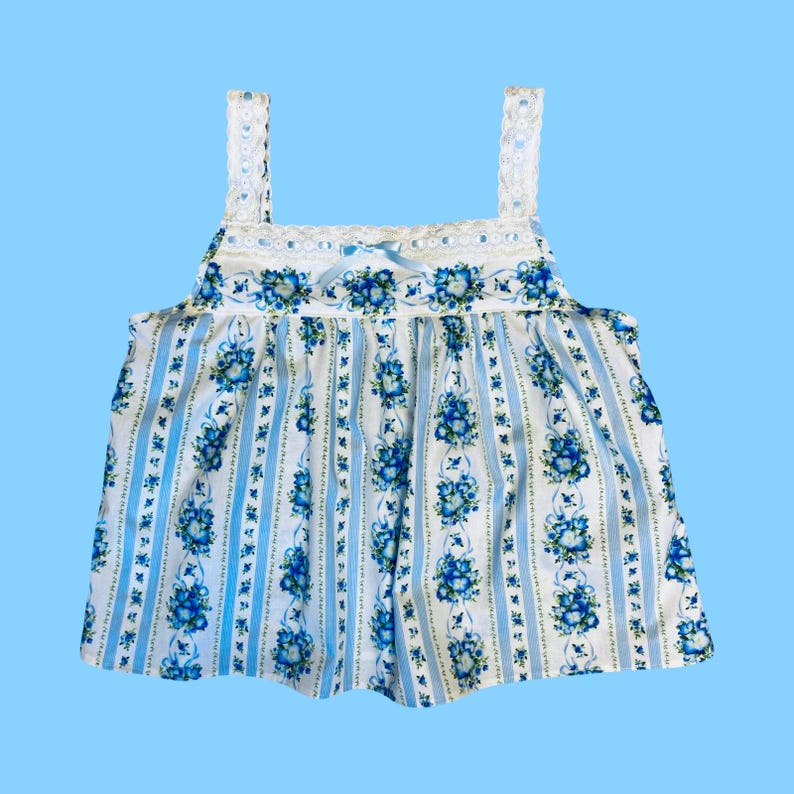 Może przedstawiać: Biało-niebieski top w kwiaty z kwadratowym dekoltem i koronkowymi ramiączkami. Top posiada wz&oacute;r niebieskich kwiat&oacute;w i pionowych niebieskich pask&oacute;w. Mała niebieska kokarda znajduje się na środku dekoltu.