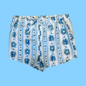 Może przedstawiać: Biało-niebieskie szorty w kwiaty ze wstążką w talii. Szorty mają pionowy wz&oacute;r w paski z niebieskimi bukietami kwiat&oacute;w. Tło jest jasnoniebieskie.