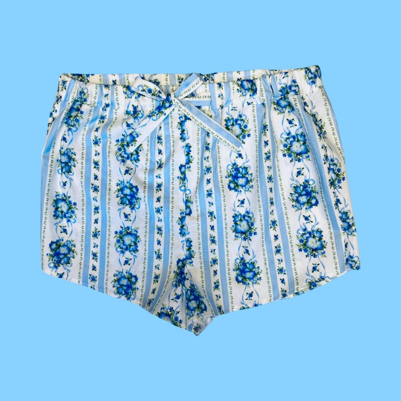 Może przedstawiać: Biało-niebieskie szorty w kwiaty ze wstążką w talii. Szorty mają pionowy wz&oacute;r w paski z niebieskimi bukietami kwiat&oacute;w. Tło jest jasnoniebieskie.