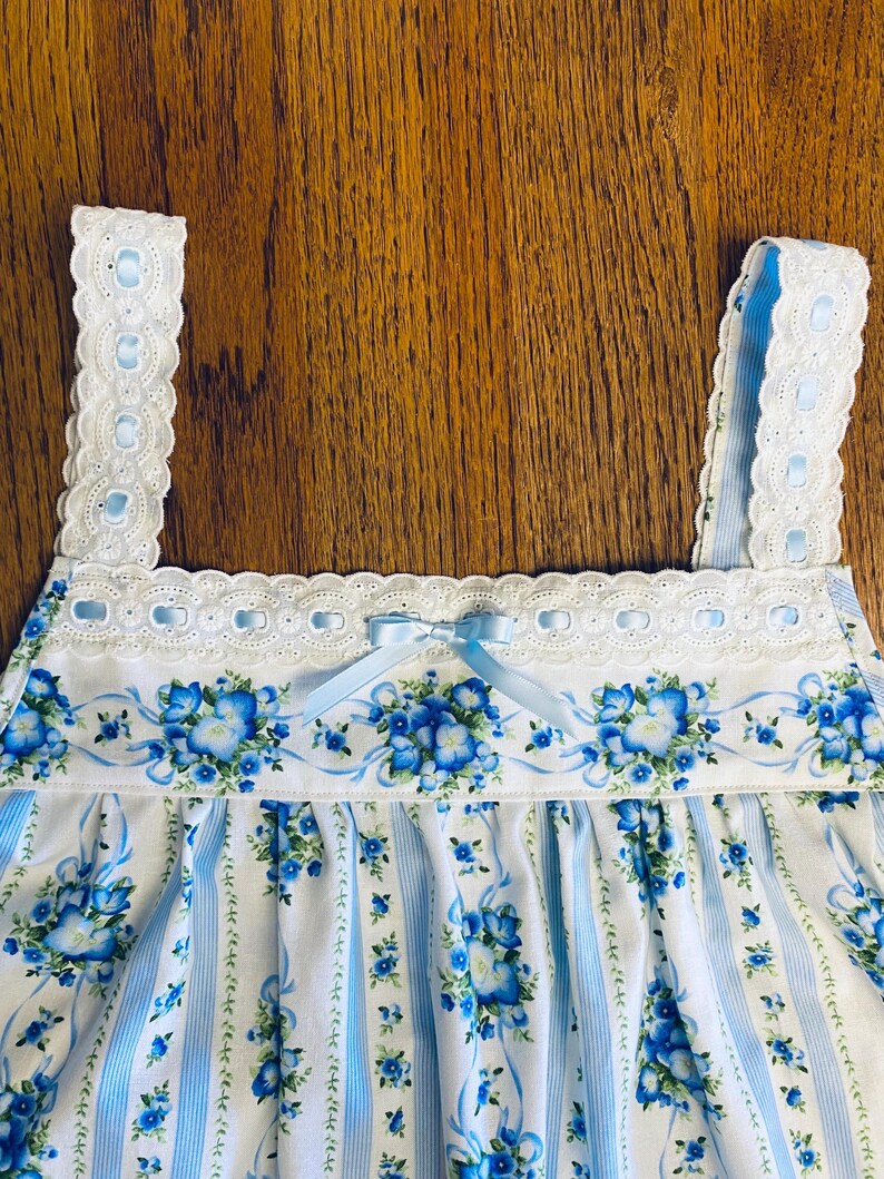 Może przedstawiać: Nocna koszula w niebiesko-biały kwiatowy wz&oacute;r z jasnoniebieską kokardą. Koszula nocna ma kwiatowy wz&oacute;r z niebieskimi kwiatami i wstążkami. Paski i dekolt są obszyte białą koronką.