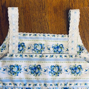 Może przedstawiać: Biało-niebieski element garderoby z kwiatowym nadrukiem i niebieskimi wstążkami. Element garderoby ma powtarzający się wz&oacute;r niebieskich kwiat&oacute;w i jest wykończony białą koronką i ramiączkami z oczkami. Tkanina ma pionowe niebieskie paski.