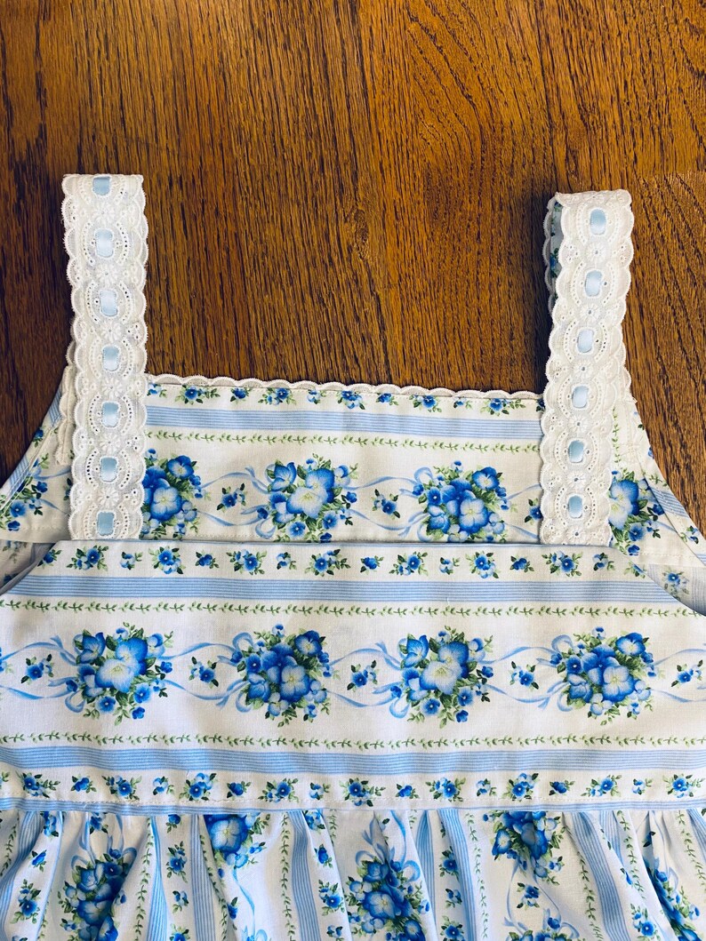 Może przedstawiać: Biało-niebieski element garderoby z kwiatowym nadrukiem i niebieskimi wstążkami. Element garderoby ma powtarzający się wz&oacute;r niebieskich kwiat&oacute;w i jest wykończony białą koronką i ramiączkami z oczkami. Tkanina ma pionowe niebieskie paski.