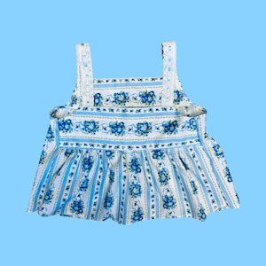 Może przedstawiać: Top w stylu vintage z niebieskim kwiatowym wzorem na białym tle. Top posiada pionowe niebieskie paski i falisty d&oacute;ł. Paski są białe z koronkowym wykończeniem. Całkowity design przypomina retro sukienkę.