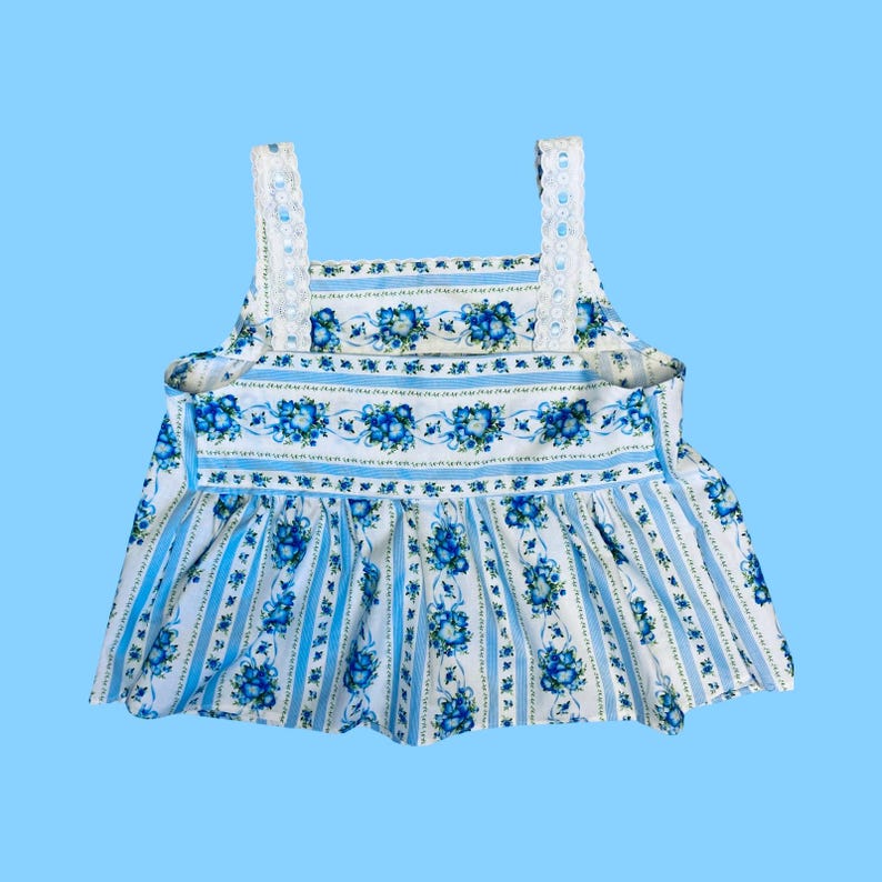 Może przedstawiać: Top w stylu vintage z niebieskim kwiatowym wzorem na białym tle. Top posiada pionowe niebieskie paski i falisty d&oacute;ł. Paski są białe z koronkowym wykończeniem. Całkowity design przypomina retro sukienkę.