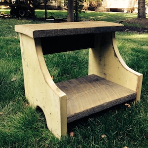 May include: A small wooden stool with a light yellow paint finish and a dark brown wood top. The stool is sitting on a green grassy lawn.