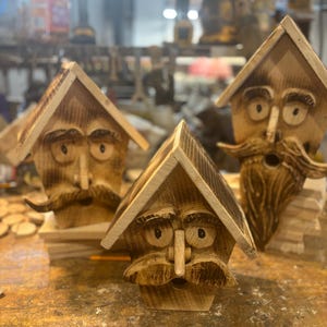 May include: Three wooden birdhouses with carved faces, mustaches, and eyebrows. The birdhouses have a natural wood finish and a triangular roof. The birdhouses are on a wooden surface.