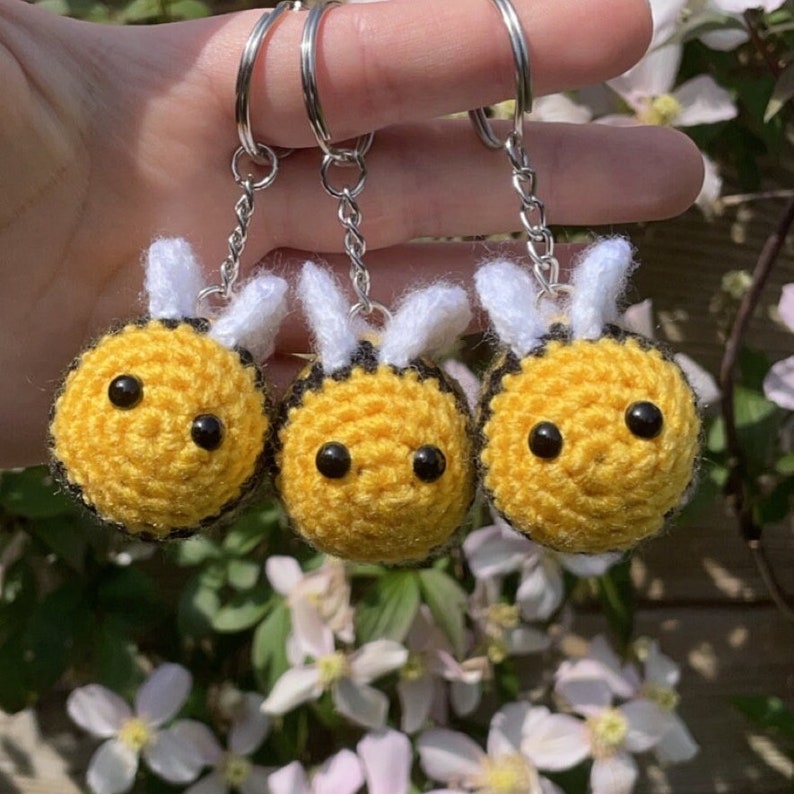 Peut inclure: Trois porte-cl&eacute;s abeilles faits &agrave; la main au crochet. Chaque abeille est jaune avec des rayures noires, des yeux noirs et des ailes blanches. Les porte-cl&eacute;s ont des anneaux et des cha&icirc;nes en m&eacute;tal argent&eacute;. Le fond pr&eacute;sente un feuillage vert et des fleurs blanches.