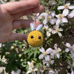 Peut inclure: Un porte-cl&eacute;s fait main en forme d'abeille crochet&eacute;e jaune et noire, avec des yeux noirs et des ailes blanches. L'abeille est attach&eacute;e &agrave; un porte-cl&eacute;s argent&eacute;. En arri&egrave;re-plan, des fleurs blanches et roses.