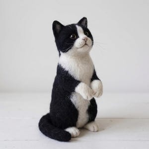 Peut inclure: Une figurine de chat en laine feutrée en position debout avec une fourrure noire et blanche. Le chat présente un poitrail, un ventre et une tache faciale blancs, avec des oreilles, un dos et une queue noirs. Il a des yeux marron foncé.