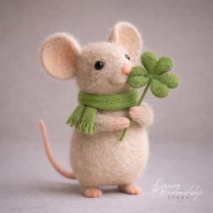 May include: A handmade felt mouse figure with a light beige body, large ears, and a pink nose and tail. It wears a green scarf and holds a green four-leaf clover. The figurine is a charming, whimsical decoration.