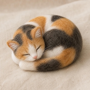Puede incluir: Una figura de gato calicó de lana afieltrada en posición de sueño. El gato presenta una cara blanca con manchas naranjas y gris oscuro en la cabeza, el lomo y la cola, con una pequeña nariz rosa y delicados bigotes blancos en el hocico.