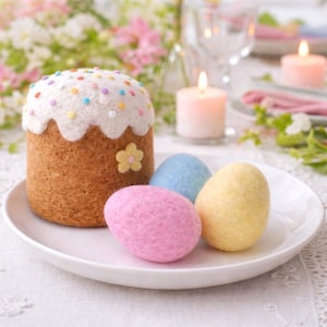 Könnte beinhalten: Ein Filz-Osterkuchen mit weißer Spitze, verziert mit bunten Streuseln und einer gelben Blume. Drei Filz-Ostereier in Rosa, Blau und Gelb liegen auf einem weißen Teller. Kerzen und Blumen im Hintergrund.