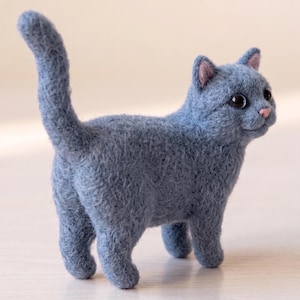 Pode incluir: Uma estatueta de gato em lã feltrada com agulha e acabamento texturizado em azul ardósia. O gato está de pé com a cauda levantada, apresentando olhos escuros e redondos, pequenas orelhas cor-de-rosa por dentro e um narizinho rosa, em pose felina.