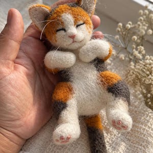 Puede incluir: Una figura de gato calicó de lana fieltrada con pelaje blanco, naranja y gris oscuro. El gato está representado en posición de dormir con los ojos cerrados, una pequeña nariz rosa y almohadillas rosas visibles en las patas. Largos bigotes blancos.