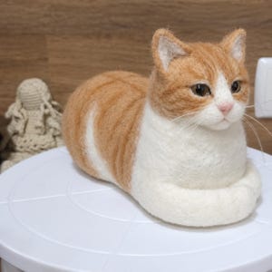 Pode incluir: Uma estatueta de gato feita em feltro de agulha com pelo laranja e branco. O gato está representado numa posição de repouso com as patas dianteiras dobradas sob o corpo. Tem olhos escuros, um nariz cor-de-rosa e longos bigodes brancos.