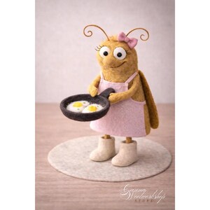Könnte beinhalten: Eine skurrile Filz-Insektenfigur mit rosa Schürze und Schleife, die eine Bratpfanne mit zwei Spiegeleiern hält. Die Figur hat große Augen, lockige Antennen und trägt cremefarbene Stiefel. Sie steht auf einem hellgrauen Filzkreis.