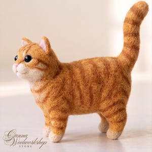 May include: A needle-felted orange tabby cat figurine with dark orange stripes, a cream-coloured muzzle, and black bead eyes. The cat stands with its tail held high, featuring light-coloured paws and pink inner ears. The text Ganna Woolworkshop Store is visible.