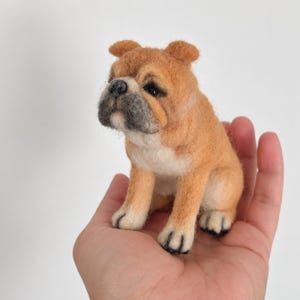 Peut inclure: Une petite figurine de bouledogue en laine feutrée, aux poils fauves et blancs. Le chien présente un museau gris foncé, une truffe noire et des yeux sombres, avec un aspect texturé et duveteux. Il est représenté assis, les pattes avant posées.