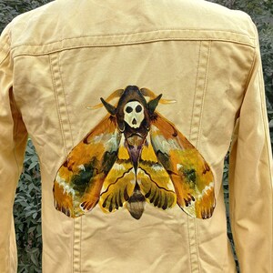 May include: Mustard yellow denim jacket featuring a large moth illustration on the back. The moth design includes a skull and wings in shades of brown, yellow, and green. The jacket has long sleeves and a classic collar.