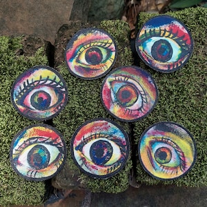 May include: Seven colorful round patches with painted eyes. Each eye is a different color combination with black pupils. The patches are made of fabric and have a slightly textured surface.