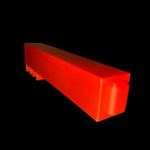 May include: A bright red rectangular object with a series of small, triangular notches along one side. The object has a smooth, glossy finish and a rounded end. The background is solid black, highlighting the object's shape and colour.