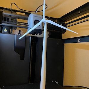 Puede incluir: Modelo blanco impreso en 3D de un aerogenerador. El modelo presenta una torre alta y cilíndrica que sostiene tres aspas. Las aspas están conectadas a un cubo central. El modelo está sobre una superficie negra.