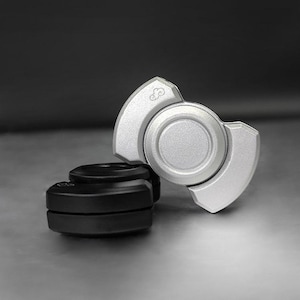 May include: A silver fidget spinner with a three-lobed design, resting on a black base. The spinner has a circular center and curved arms. The background is a gradient of gray.
