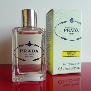 Könnte beinhalten: Ein rechteckiger Prada-Parfümflakon mit silbernem Verschluss und passender Schachtel. Flakon und Schachtel tragen das Prada-Logo und die Aufschrift "Milano" und "Infusion D'Ylang". Der Flakon enthält 7,5 ml Parfüm.