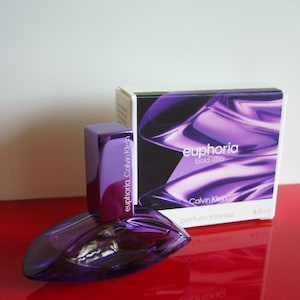 Può includere: Un set di bottiglia e scatola di profumo viola. La bottiglia è di forma ovale con un tappo rettangolare abbinato. La scatola presenta le parole "euphoria bold elixir" e "Calvin Klein" in testo bianco su sfondo viola. Il set è su una superficie rossa.
