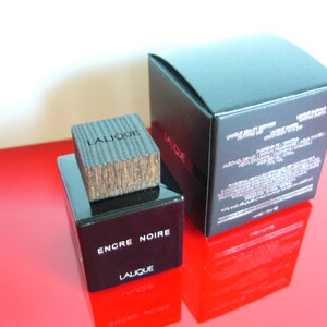 Peut inclure: Un flacon de parfum noir rectangulaire avec un bouchon en bois et les mots "ENCRE NOIRE" et "LALIQUE" imprim&eacute;s sur le devant. Une bo&icirc;te noire avec du texte blanc est derri&egrave;re le flacon. Le flacon et la bo&icirc;te sont sur une surface rouge.