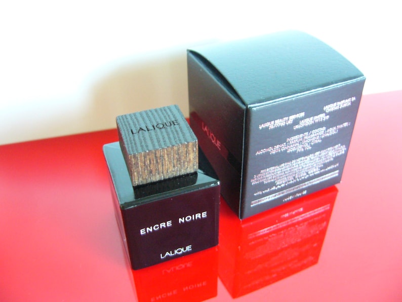 Peut inclure: Un flacon de parfum noir rectangulaire avec un bouchon en bois et les mots "ENCRE NOIRE" et "LALIQUE" imprim&eacute;s sur le devant. Une bo&icirc;te noire avec du texte blanc est derri&egrave;re le flacon. Le flacon et la bo&icirc;te sont sur une surface rouge.
