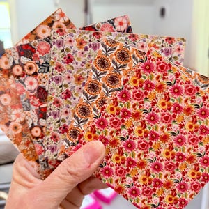 May include: A hand holds a fan of floral patterned paper sheets. The sheets display a variety of colorful floral designs in shades of red, orange, pink, and purple. The patterns include detailed flower and leaf arrangements.