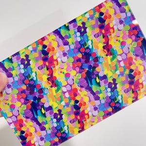 May include: A rectangular piece with a vibrant, colorful abstract design. The artwork features a textured pattern of small, overlapping dots in shades of purple, pink, yellow, green, and blue. The piece is held by a hand against a white background.