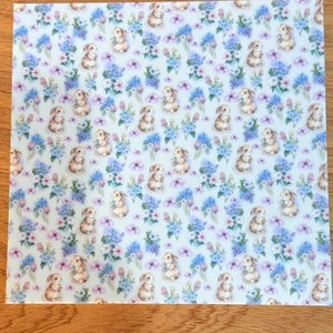May include: A square pattern with a repeating design of small, tan bunnies and blue and purple floral arrangements on a pale blue background. The design has a soft, spring-like appearance.