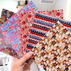May include: A hand holds a fan of patterned sheets. The sheets display various designs, including floral, patriotic stars and stripes, and a repeating pattern of cats. The color palette includes red, blue, white, and a variety of other colors.