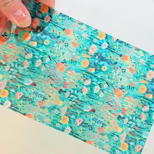 May include: A floral patterned paper with a teal background. The design features orange, yellow, and pink flowers with green leaves and stems. The paper is held by a hand, showcasing the vibrant colors and intricate details of the botanical print.