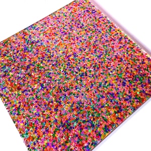 May include: A square, clear surface filled with colorful star-shaped glitter. The glitter is a mix of pink, orange, yellow, green, blue, and purple. The glitter is densely packed, creating a vibrant and festive appearance.