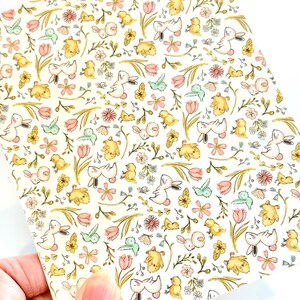 May include: A sheet of patterned paper with a repeating design of bunnies, chicks, flowers, and butterflies in pastel colours. The background is off-white, and the illustrations are in shades of yellow, pink, and green.