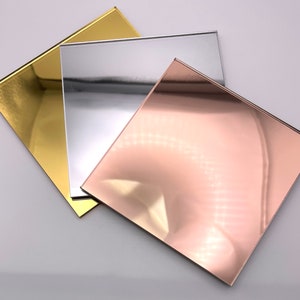 May include: Three square mirrored acrylic sheets in gold, silver, and rose gold. The reflective surfaces create a modern and elegant aesthetic. The sheets are arranged at different angles, showcasing their metallic sheen.