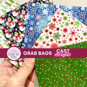 May include: A hand holds a stack of colorful acrylic sheets with festive holiday patterns. Designs include snowmen, snowflakes, Christmas trees, and string lights. The text "GRAB BAGS" and "CAST Acrylic" are visible.