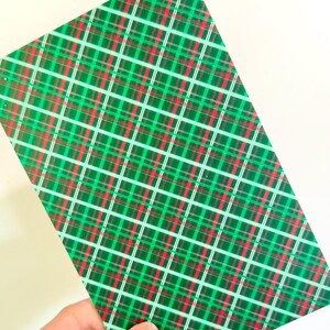 May include: A rectangular sheet with a green, red, and white plaid pattern. The plaid design features intersecting lines in a diagonal arrangement, creating a festive and decorative appearance. The sheet is held by a hand.